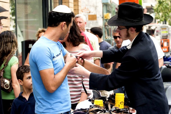 An ultra-Orthodox Jewish man helps a non religious Jewish man to fulfill the commandment in Deuteronomy 6:8 by putting on tefillin (phylacteries) for prayer. tefillin-morning prayer-Rabbi-passerby