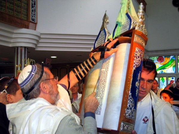 A Jewish man wearing tefillin (phylacteries) and a tallit (prayer shawl) reads from an open Torah scroll housed in an ornate Torah tik. A Jewish man wearing tefillin (phylacteries) and a tallit (prayer shawl) reads from an open Torah scroll housed in an ornate Torah tik.